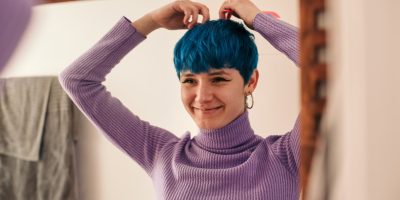 Young and natural woman with blue short hair, styling her look in front of a mirror at home, smiling and wearing a purple turtleneck jumper.