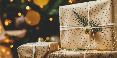 Stylish christmas gifts at christmas tree with golden lights bokeh. Merry Christmas! Wrapped christmas presents with golden paper and fir branch under decorated tree in room. Atmospheric banner